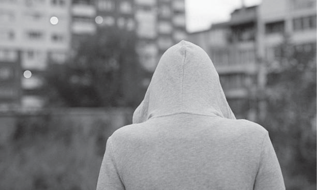 Person in a hoodie facing away, with blurred buildings in the background. Black and white image.